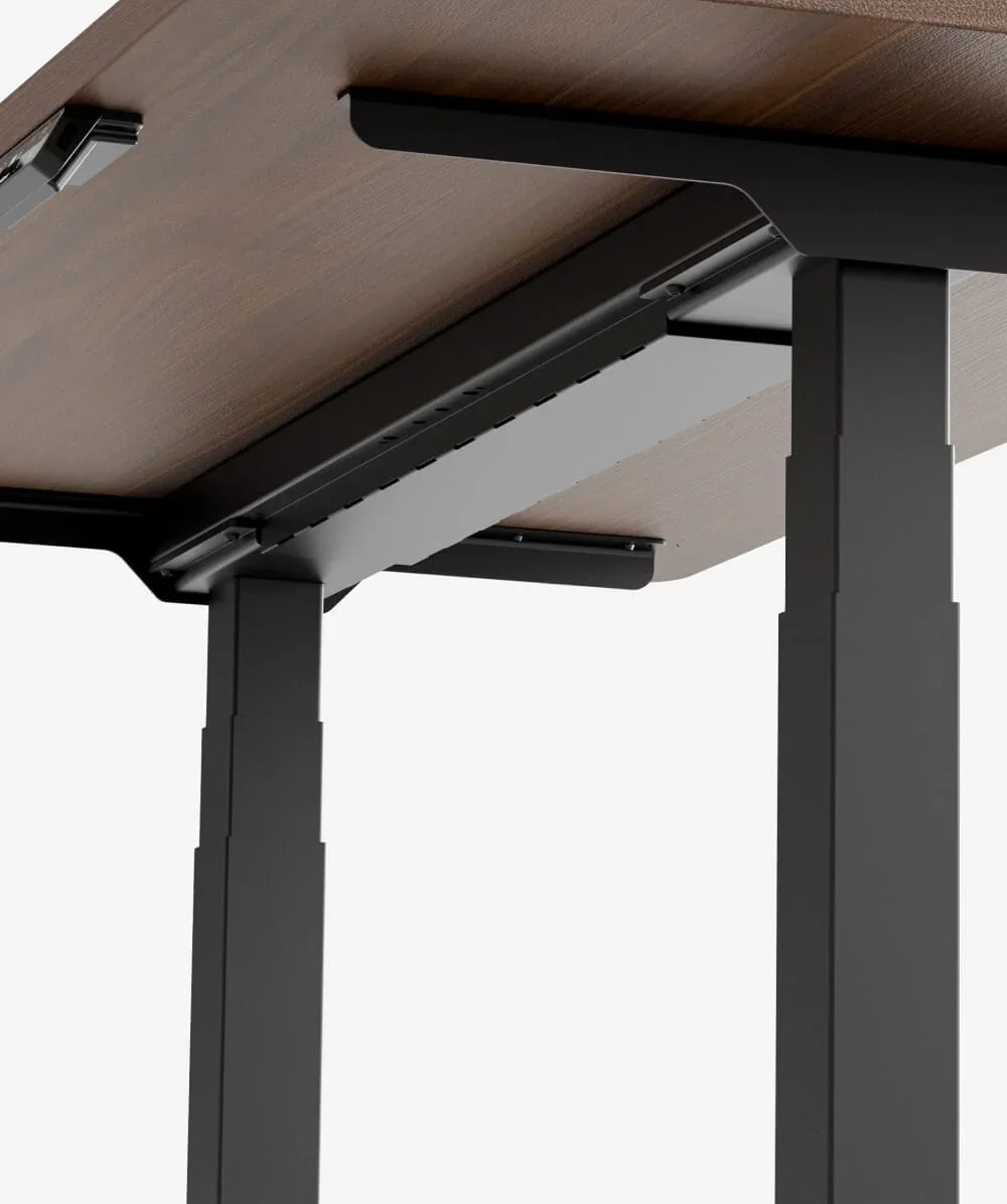 Underside close-up of a walnut top on a black C-frame showing the rear-offset legs, crossbeam and under-desk cable tray.