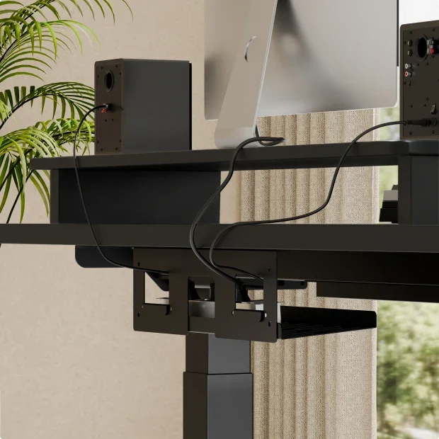 The under-desk cable tray and clamp routing cables beneath a black top, with a speaker and plant; no text.