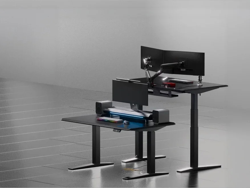 Two all-black desks in a grey studio, one lowered with a monitor, speakers and keyboard and one raised with dual monitors on an arm, illustrating the height range.