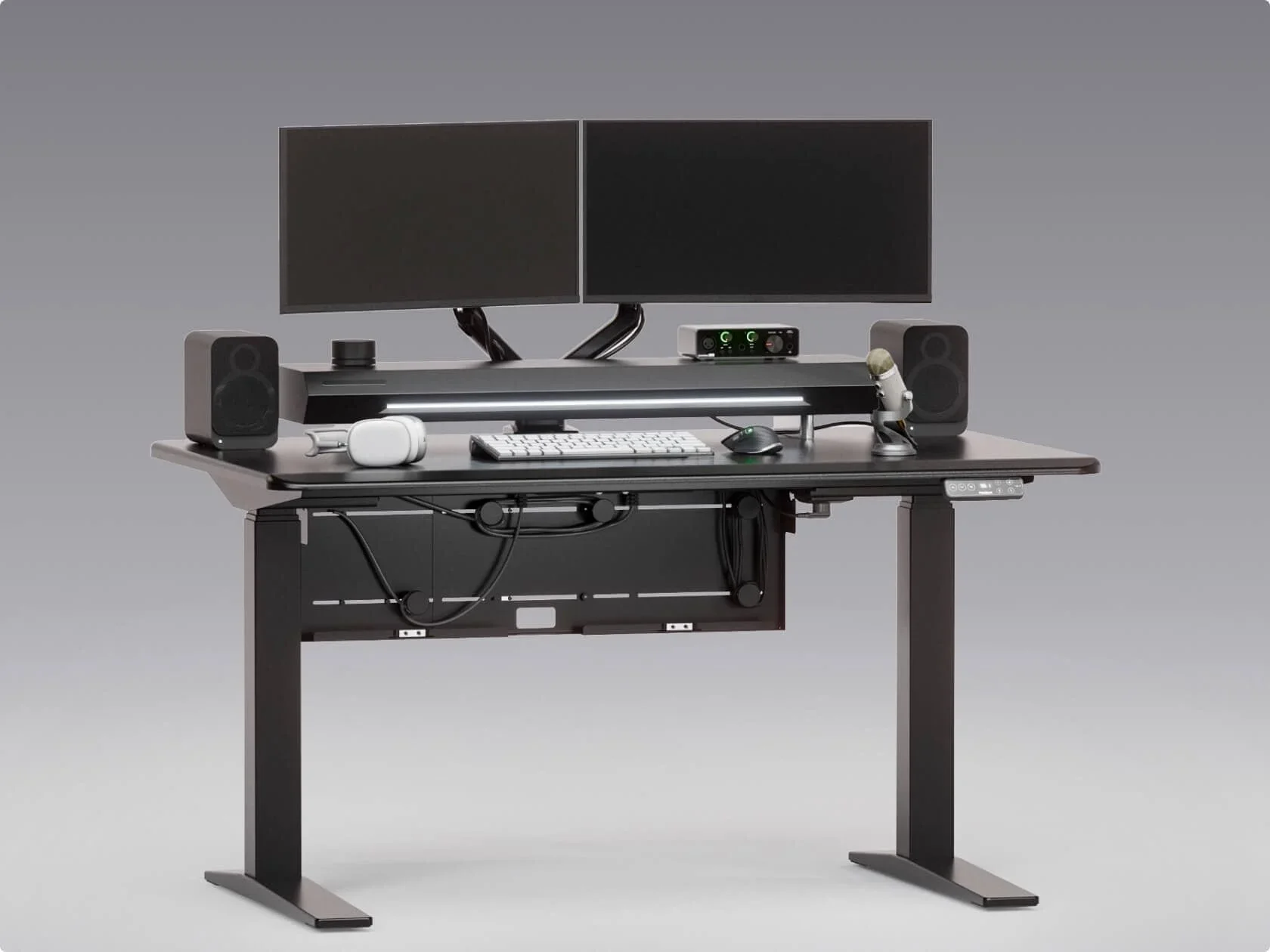 E7 Pro CableFlow electric standing desk. Studio render of the black desk with dual monitors on arms, speakers and mic; cables neatly routed into the under-desk tray (the "after" of a before/after pair).