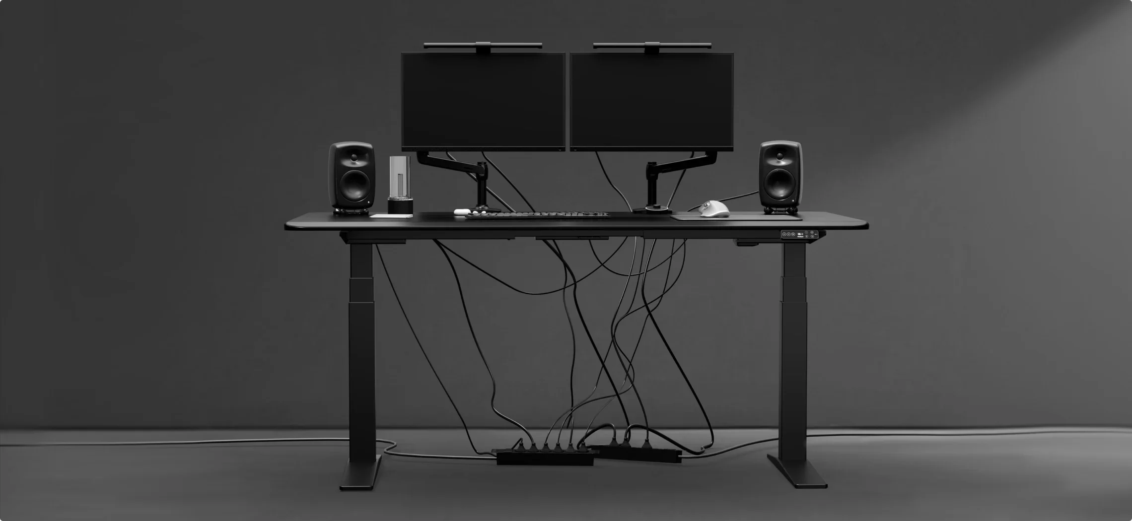 Wide render of the black desk with dual monitors and speakers against a grey wall, with loose cables hanging down to a floor power strip.