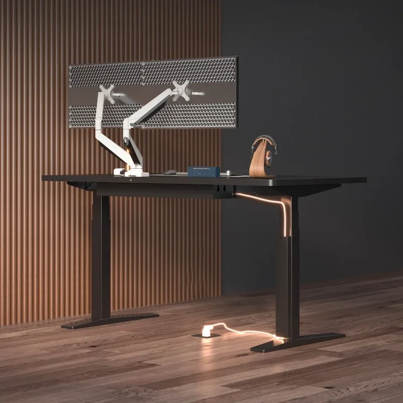 Black desk with a white dual monitor arm, headphone stand and small devices in a warm wood-slat room, with a lit cable running down the leg to a floor socket.