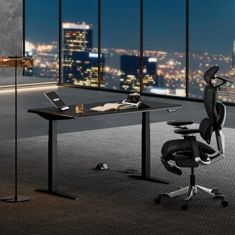 All-black desk in a dark high-rise office at night with city lights through floor-to-ceiling windows, a tablet and open book on the desk and a black mesh ergonomic chair beside it.