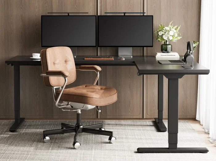 Black E7L-Lite L-desk with dual monitors, keyboard and flowers, with a tan leather office chair, in a room with wood-panelled walls and sheer curtains.
