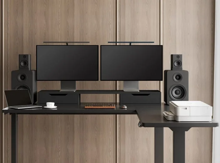 Front view of a black E7L-Lite L-desk with dual monitors on a riser, bookshelf speakers, a printer and laptop against wood-panelled walls.