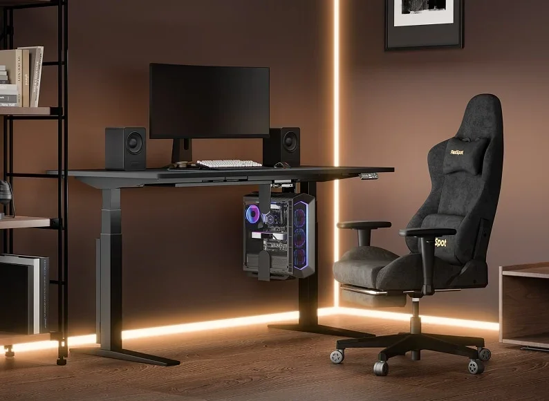 Black desk in a brown-walled gaming corner with an LED strip, a monitor, speakers, a PC hung under the desk, shelving and a black fabric gaming chair.