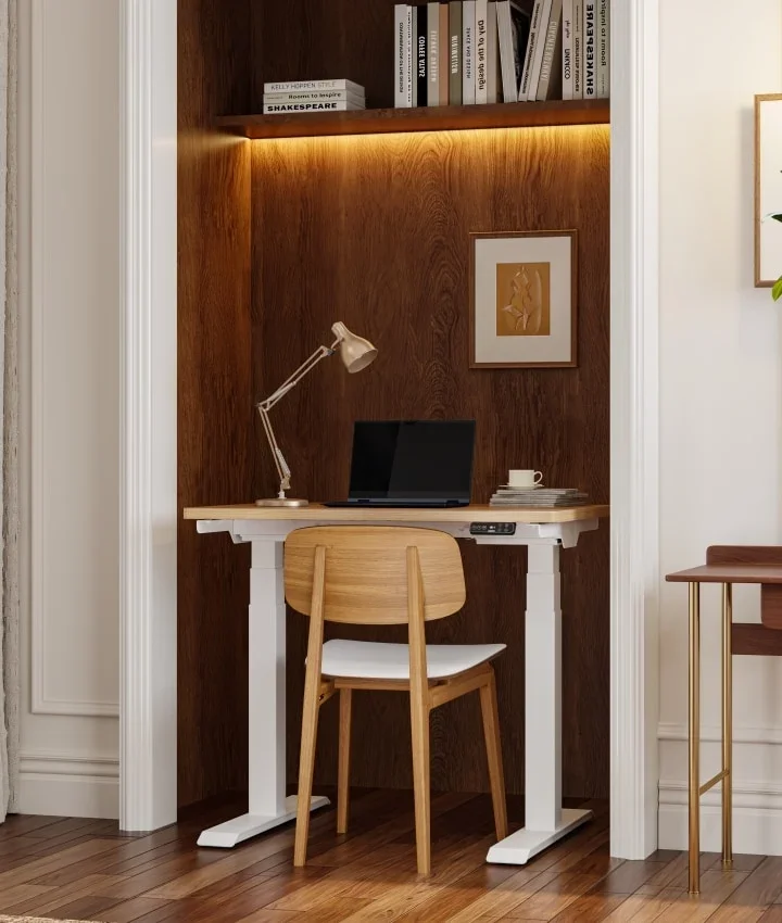 White-frame compact desk with an oak top, laptop and lamp tucked into a dark wood alcove with shelves.